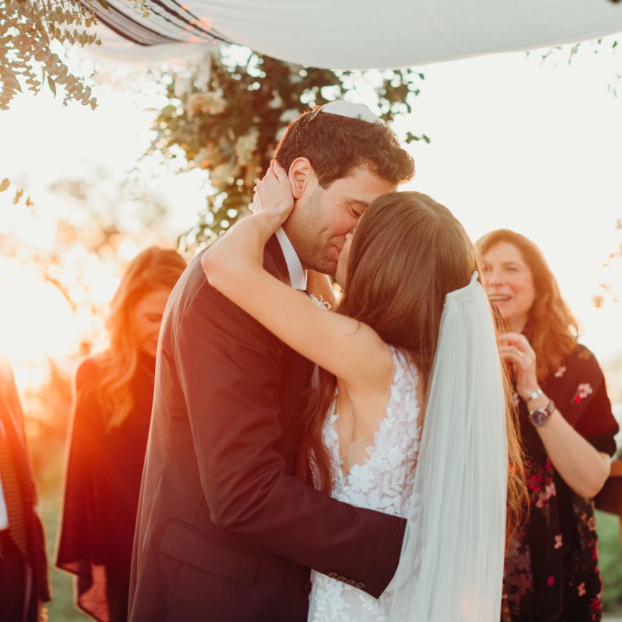 Bride and groom kissing at sunset beneath a chuppah at an outdoor ceremony