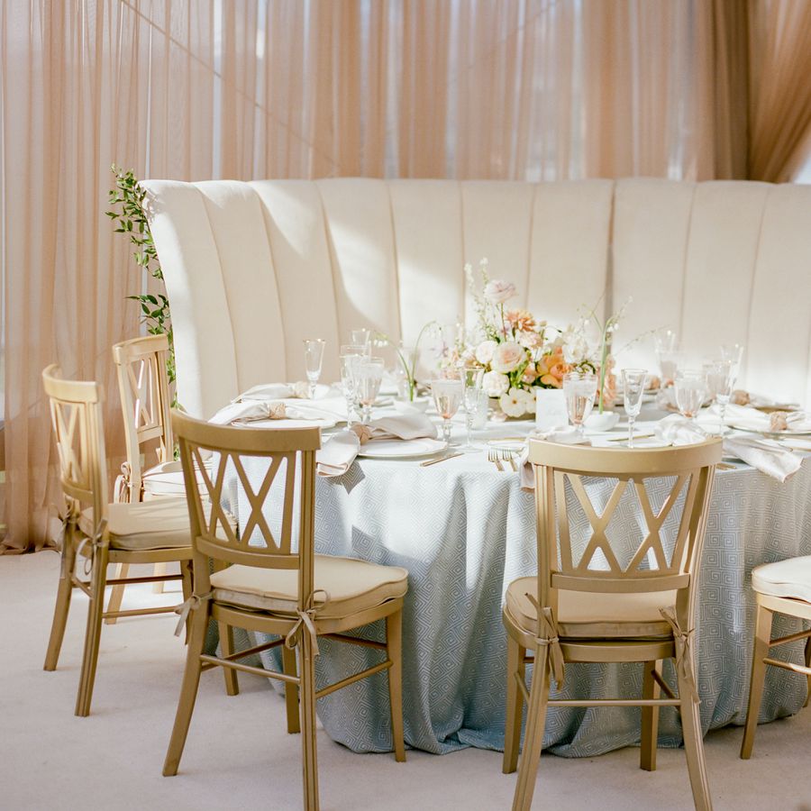 A round wedding reception table with white table cloth, pastel flowers, glassware, wooden chairs, and upholstered high-back booth seating.