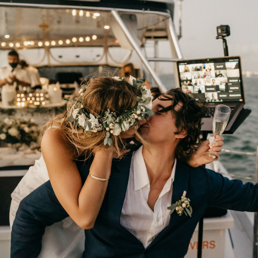 Bride kissing groom while holding champagne on a boat while guests watch from a live-stream app on a laptop