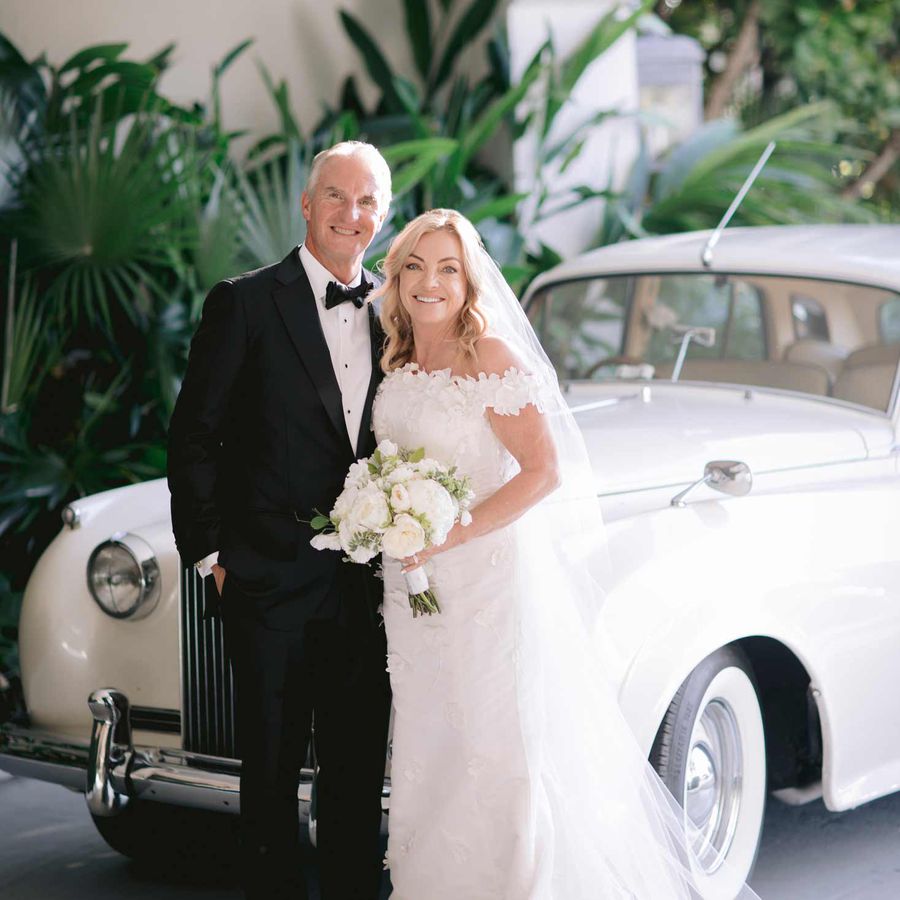 Sean Rooney in black tuxedo and Colleen Rooney in white off-the-shoulder gown holding white bouquet in front of a white vintage car