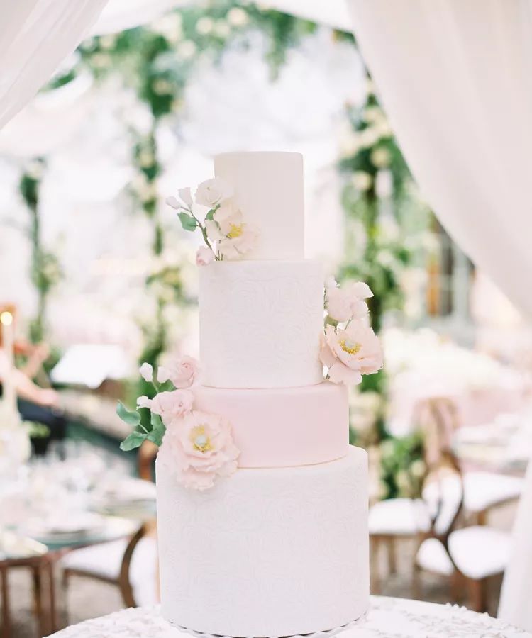 A four-tiered pink and white wedding cake covered in flowers at a tented wedding reception.