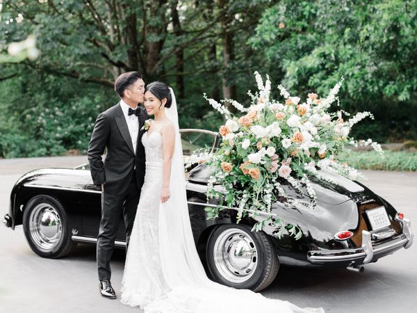 bride and groom posing in front a black vintage getaway car, which is decorated with white, orange, and pink roses