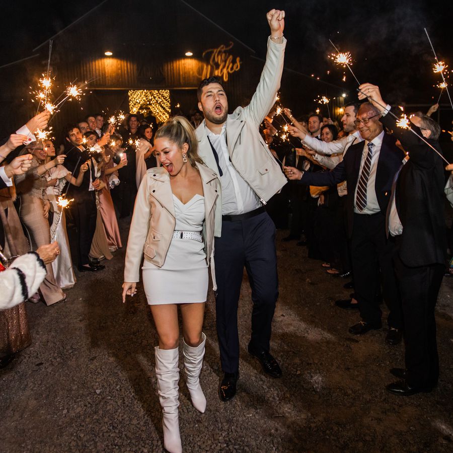 Bride and groom celebrating while guests wave sparklers in the air as they leave their barn reception venue