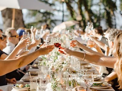 An outdoor wedding reception with guests seated at a long dinner table with flowers and candles, raising wine glasses
